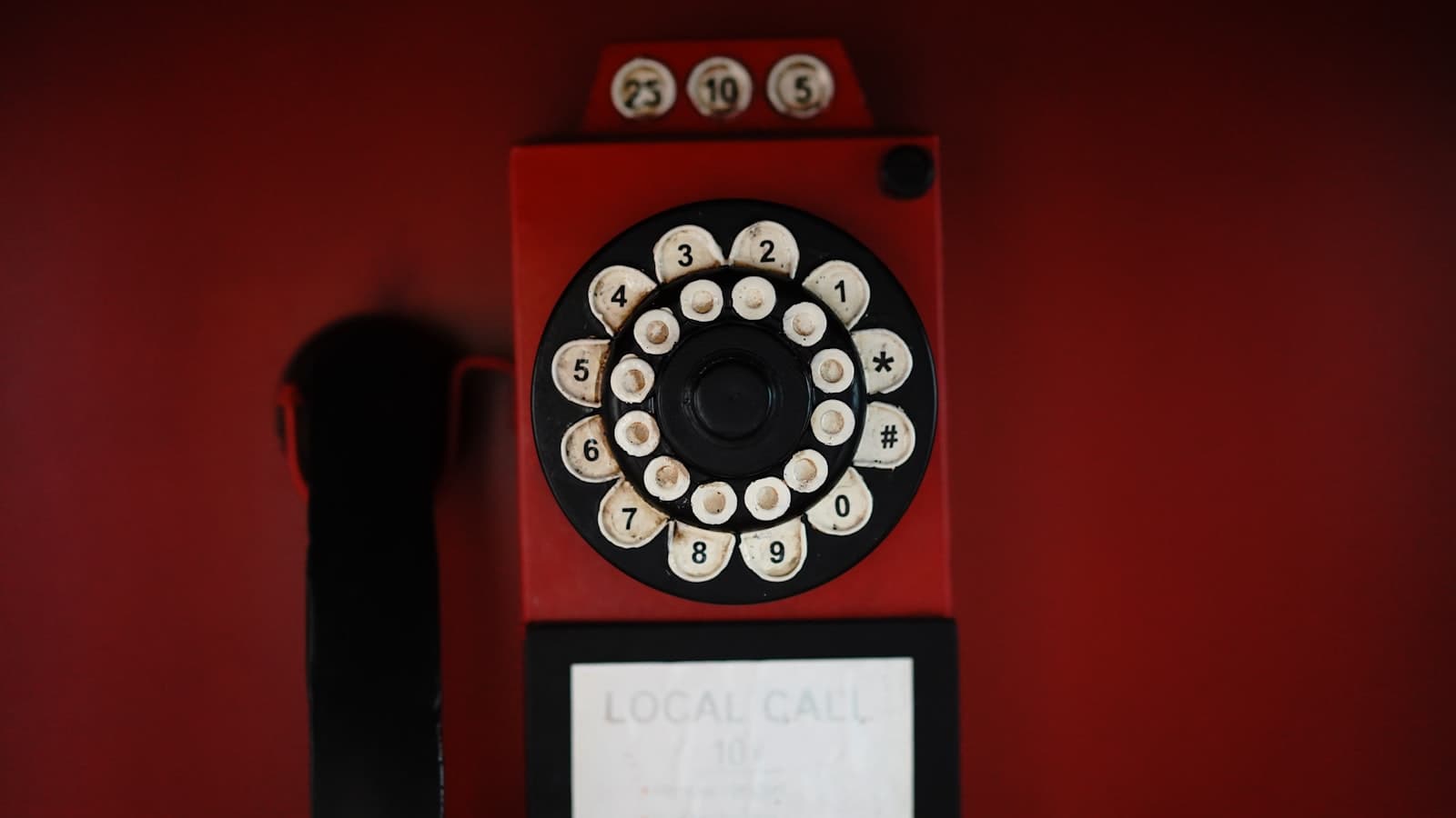 Editorial photograph of a vintage red payphone with a worn rotary dial against a deep red wall.