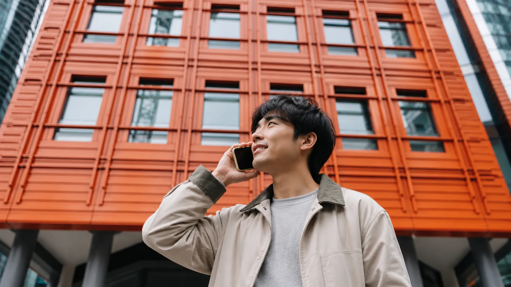 A young man standing outside a modern office building, smiling as he takes a call on his smartphone.